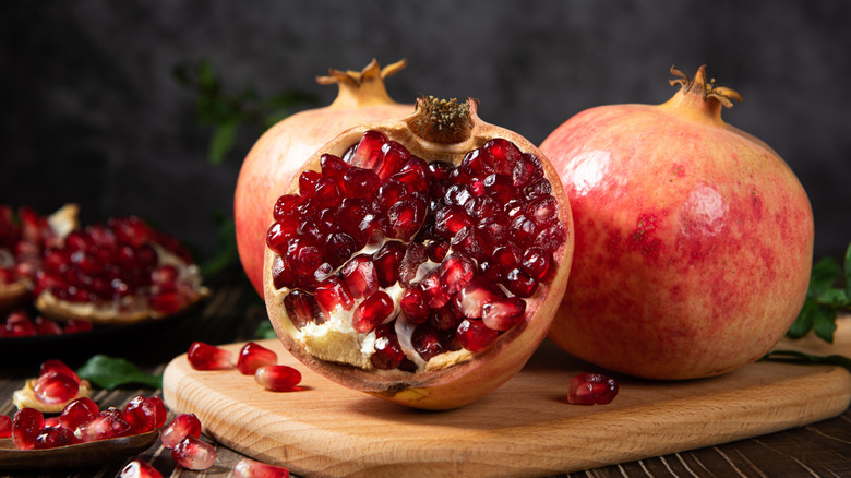 A halved pomegranate spilling seeds out onto a board with two whole pomegranates behind it
