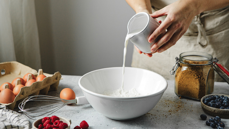 Hands pouring milk into mixing bowl on countertop, surrounded by other baking ingredients