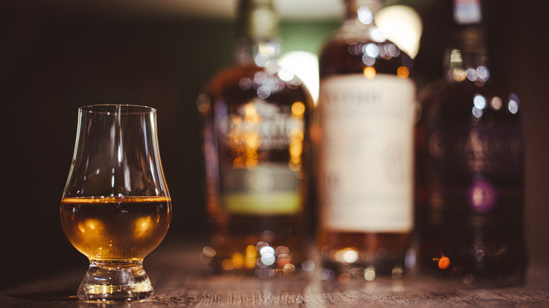 A filled glass of whiskey placed on a table with blurred bottles in the background