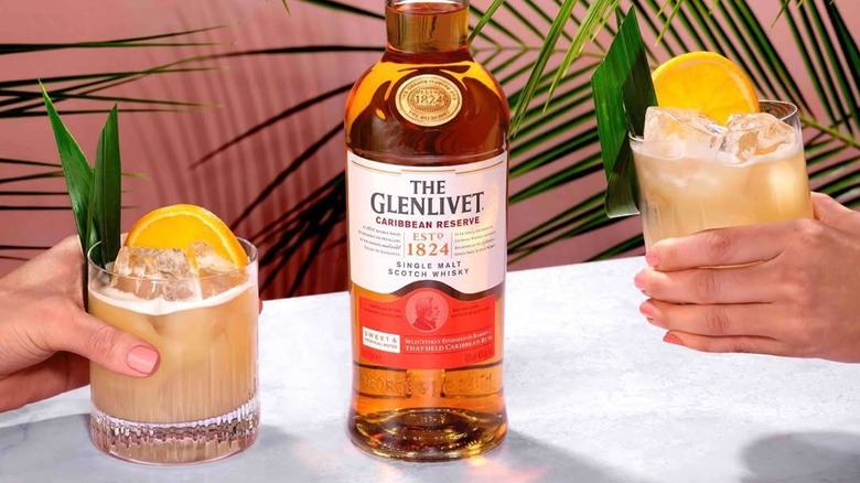 A bottle of The Glenlivet Caribbean Reserve with people holding tropical cocktails either side