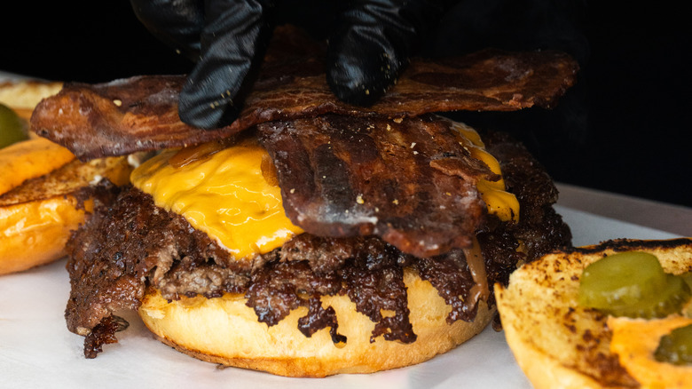 A smash burger with a  gloved hand putting bacon on top of the patties