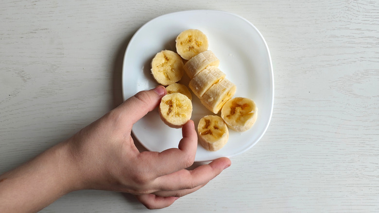 Hand holding banana slice above white plate with several banana slices on it