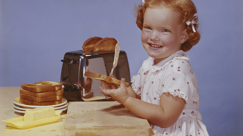 1960s-style photo of young girl holding a slice of toast at the table, next to a toaster and a pile of toast and a slab of butter