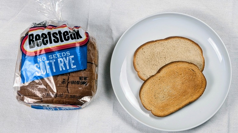 A loaf of Beefsteak No Seeds rye bread next to a white plate with two slices of bread on a white tablecloth