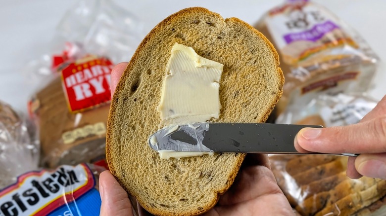 A close-up of a hand using a butter knife to smear butter on a slice of rye toast with loaves in plastic packages on a table in the background