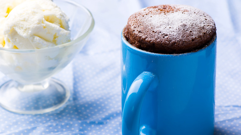 a tall, fluffy chocolate mug cake in a vivid blue mug, on a pale blue tablecloth with white polka dots, with a coupe dish of vanilla ice cream to its left