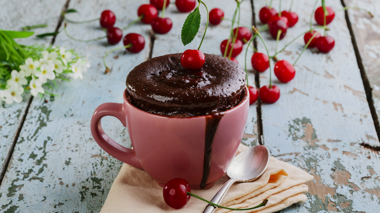chocolate cake in a pink mug, shown on a rustic (painted, distressed) wooden surface, with a spoon and folded napkin, garnished with a fresh cherry, with a scattering of additional cherries and a small spray of white flowers visible in the background