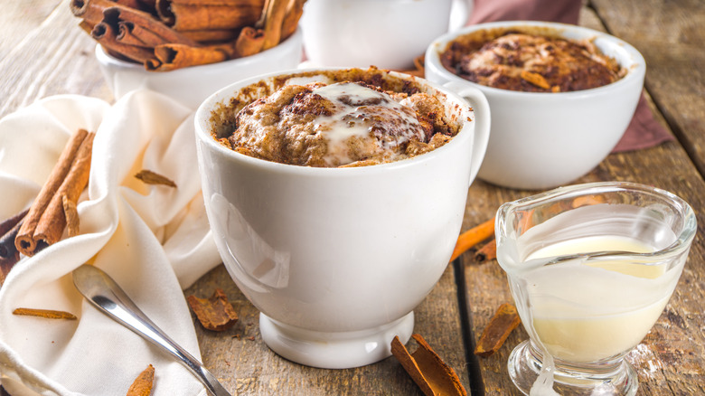 a cinnamon-flavored coffee cake in a white mug, pictured on a rustic surface with a spoon, a thin, custardy sauce, and whole and broken cinnamon sticks