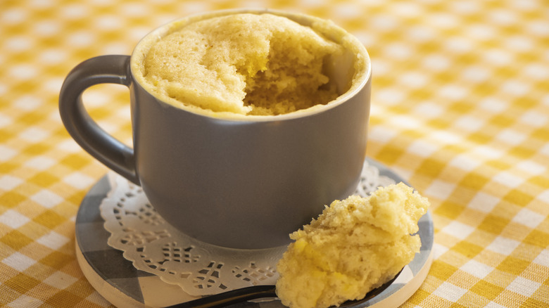 yellow cake in a grey coffee mug, with a single spoonful of cake sitting at its side on a doily, against the background of a yellow-checked tablecloth
