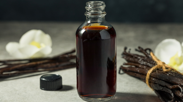 bottle of natural vanilla extract on a pale grey counter with a dark background; cap of the bottle is beside it, and it's flanked by two bundles of vanilla beans, each with a vanilla blossom behind it