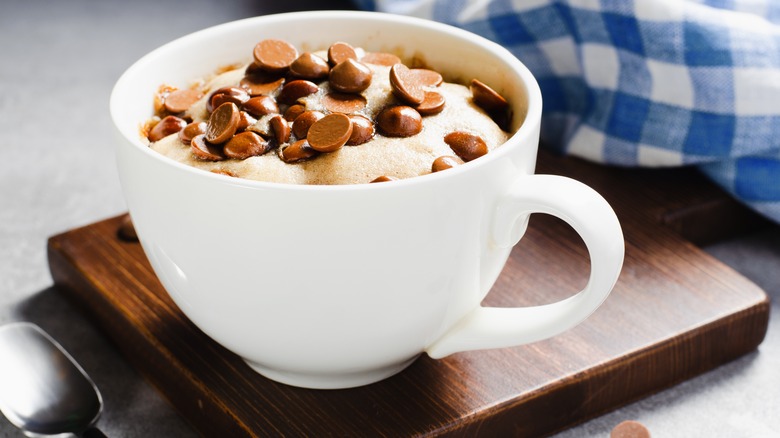 peanut butter and chocolate chip mug cake, shown in a white mug on a dark wooden trivet, with a spoon in the left foreground and a blue gingham kitchen towel in the background at top right