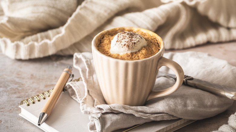 pumpkin spice mug cake topped with a dollop of whipped cream and sprinkled spices, served in a fluted white mug, shown resting on a notebook with a kitchen towel beneath it, a spoon to the rear, and a comfortable-looking off-white sweater resting rumpled in the background