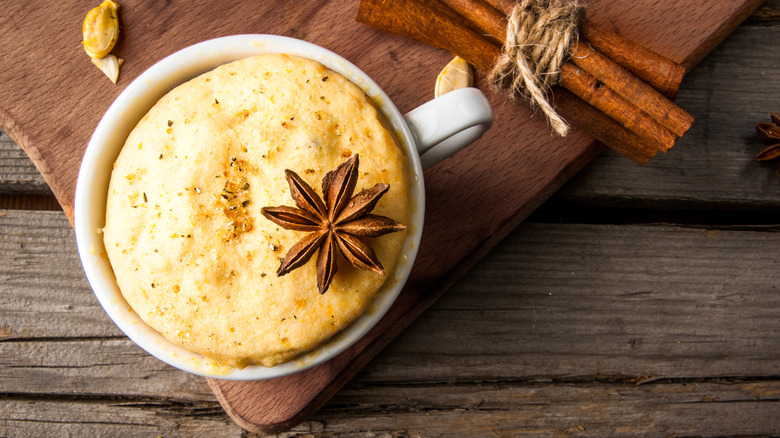spice cake in a white mug, shown in an overhead view on the corner of a cutting board, with ground spices visible on its surface as well as a star anise garnish, and a bundle of cinnamon sticks visible on the board behind it