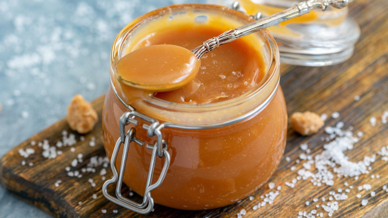 Jar of caramel sauce with spoon, on wooden board sprinkled with sea salt