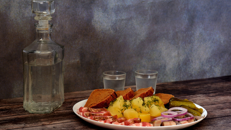 A glass decanter and two glasses of vodka on a dark wooden table and a plate with an assortment of snacks. Close-up.