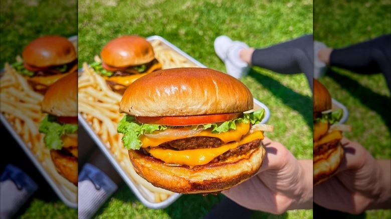 hand holding burger in front of tray with burger and fries on grass mirrored image