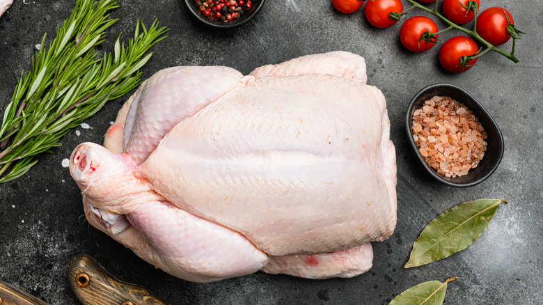 Raw whole chicken on stone surface with rosemary, bay leaves, pink salt, and cherry tomatoes.