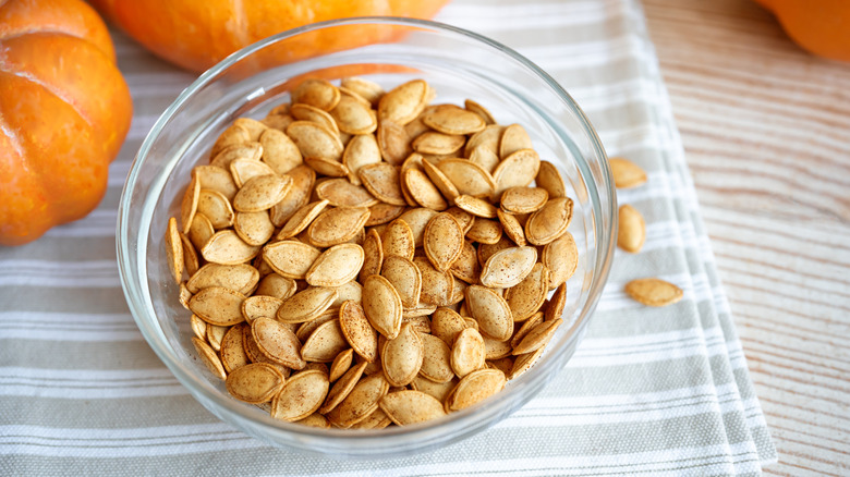 Bowl of roasted pumpkin seeds on grey striped linen, next to whole pumpkins