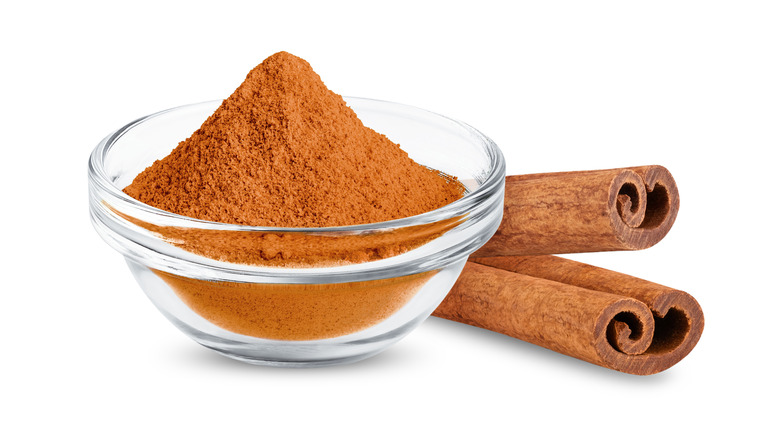 Cinnamon powder in a clear bowl and sticks on white background