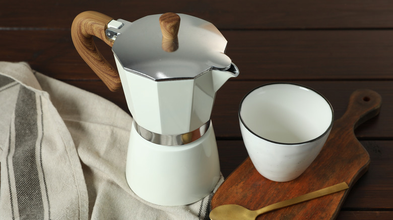 White Moka pot on towel beside white cup on cutting board with gold spoon