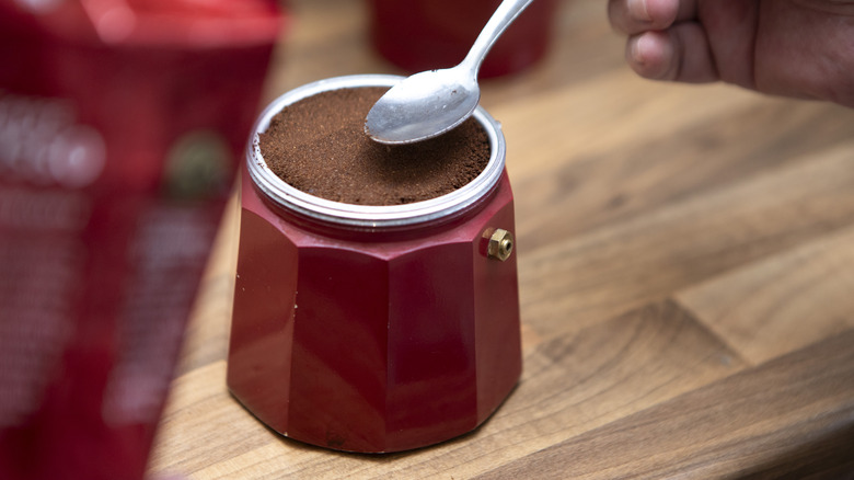 Spoon distributing coffee grounds evenly in bottom half of red Moka pot