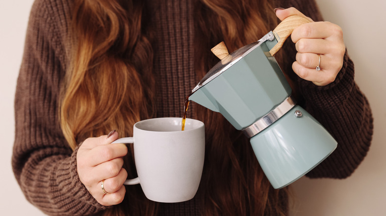 Woman pouring coffee from light blue Moka pot into white mug