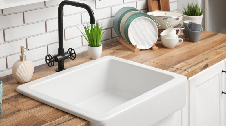 Farmhouse sink with black faucet and white brick-like backsplash next to plates, cups, and soap dispenser