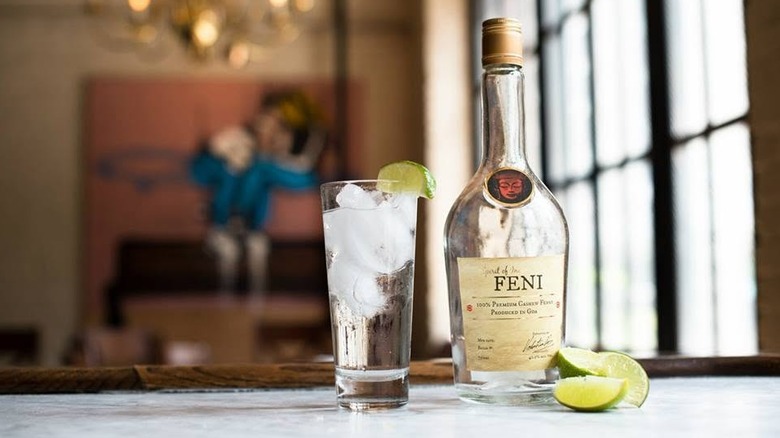 A clear bottle of feni on a table next to a highball with lime wedges on glass and scattered around.