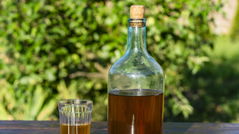 Large glass bottle and a faceted glass with homemade wine on wooden table against summer garden background, close up, outdoors.