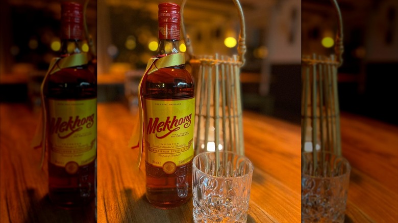 A bottle of golden Mekhong spirit against white background. Bottle has yellow label with red font and a yellow ribbon around the neck.