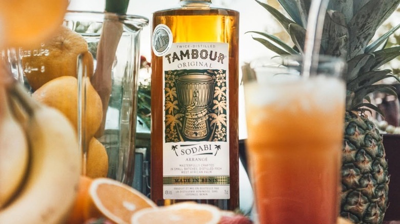 A bottle of Tambour Original around blender with citrus and pineapple, with a cocktail in the foreground.