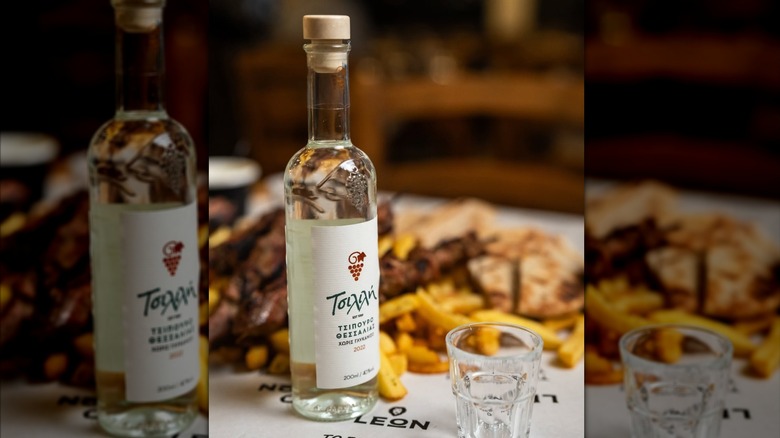 Mirror image of a small tsipouro bottle with Greek label on table beside a shot glass with botanicals in the background.