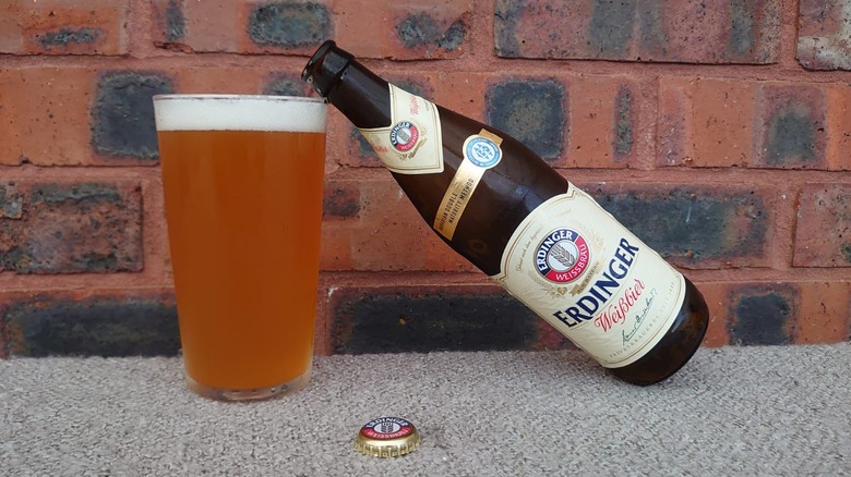 A bottle of Erdinger resting on a full glass of the beer with the bottle cap also shown