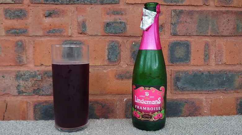 An opened bottle of Lindemans Framboise next to a glass full of the beer on a plain brick background