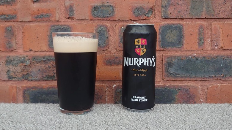 A full glass of Murphy's Irish Stout next to an opened can on a plain brick background
