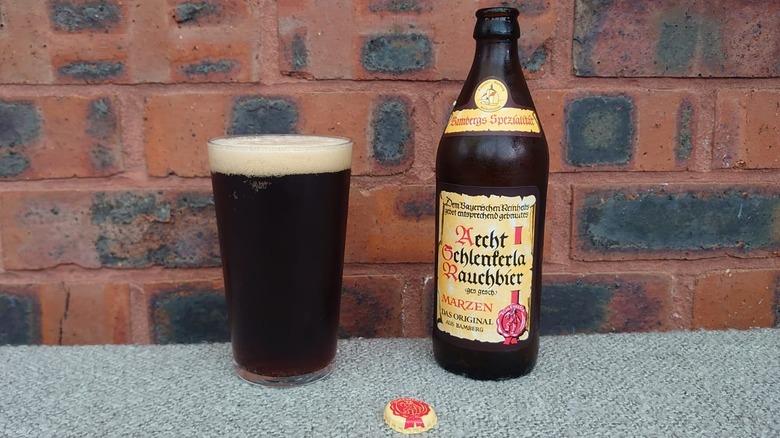 A bottle of Schlenkerla Weizen next to a glass full of the beer with a brick background and the bottle cap shown