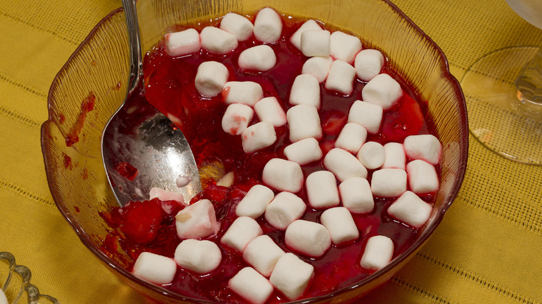 A clear bowl of strawberry and bananas and jello topped with marshmellows with spoon