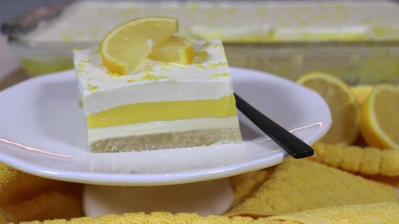 Slice of Lemon Lush on white cake dish with yellow table cloth and sliced lemons