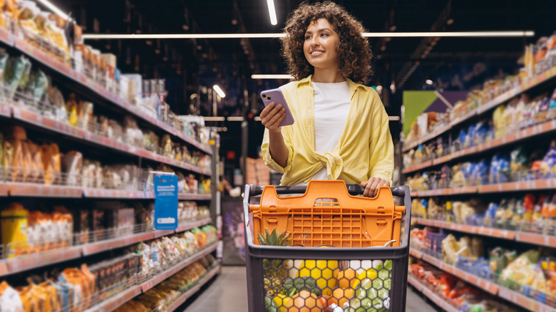 Person pushing shopping cart in grocery store while holding phone