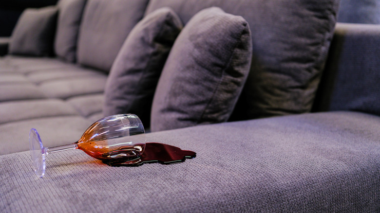 A glass of red wine is knocked over on a gray sofa.