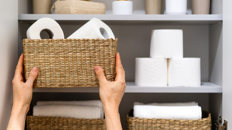 A person is placing a wicker basket containing toilet paper on a shelf in a bathroom cabinet. Woman holding box with tissue in rolls, cleaning space in tidy and neat apartment
