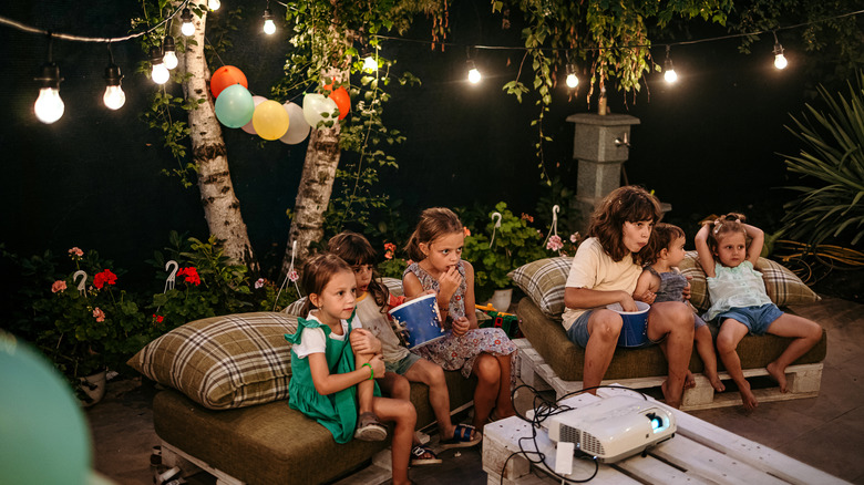 Group of children are sitting in the yard during a dinner party and watching a movie on a video projector