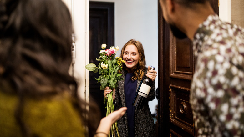 Young woman holding bouquet and wine bottle while visiting friends for dinner party.