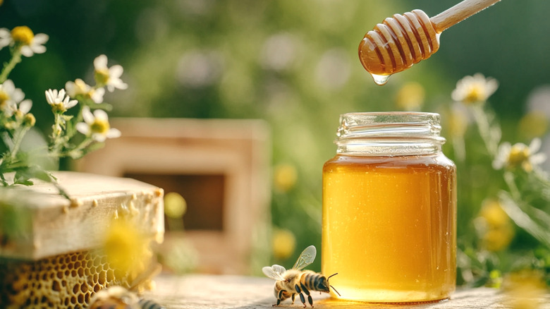 A glass jar of honey with a bee and flowers next to it