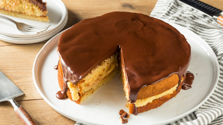 Boston cream pie  covered in a glossy chocolate ganache on white plate with a slice cut out of it