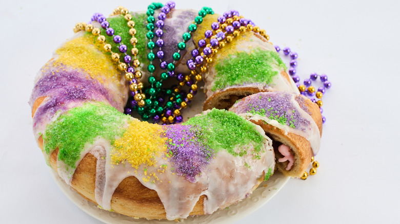 A colorful king cake draped in Mardi Gras beads and with a plastic baby peeking out from a cut slice