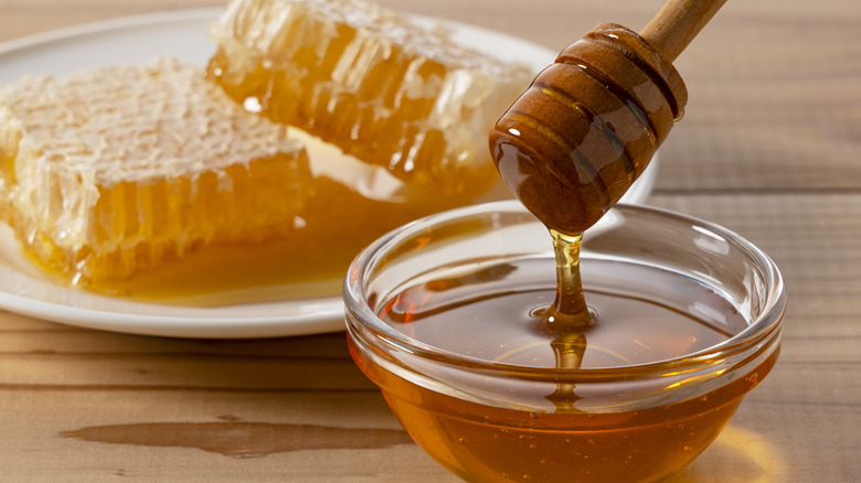 Organic Honey Dripping from Wooden Dipper into Glass Bowl, honeycomb in the background