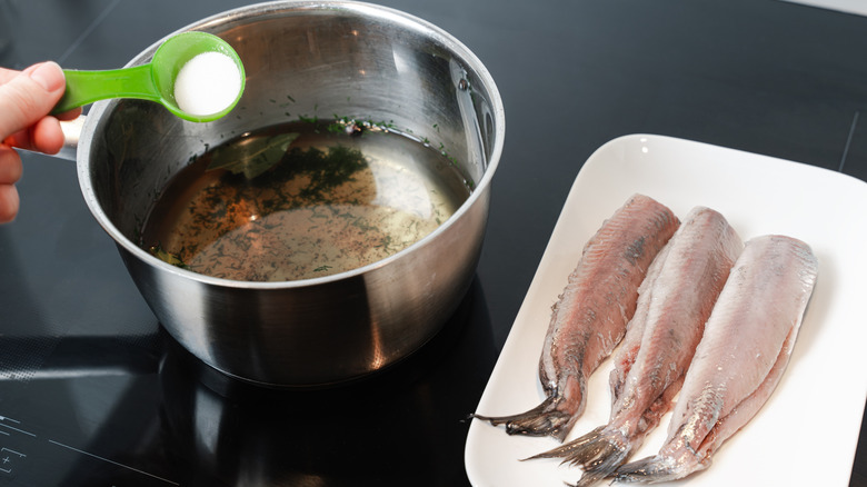 hand measuring salt into a stainless steel pot of brine, with three skinned fish fillets on a white plate