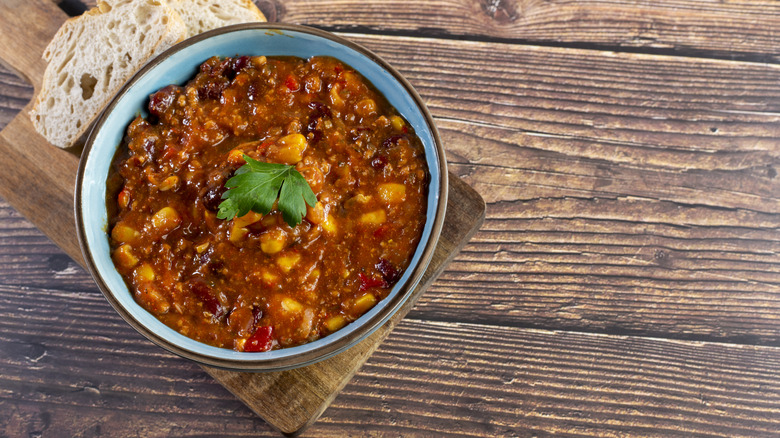 bowl of chili sitting with a slice of bread on a wooden cutting board