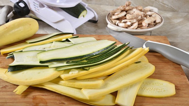wooden cutting board piled with zucchini and spaghetti squash sliced lengthwise with a bowl of mushrooms and a mandolin in the background